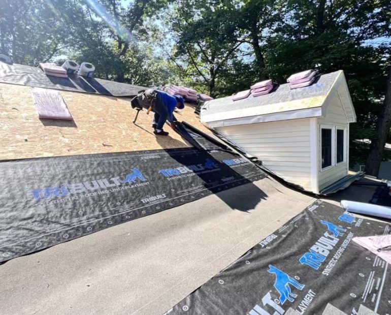 Professional roofing contractor installing synthetic roofing underlayment on residential house roof during roof replacement project