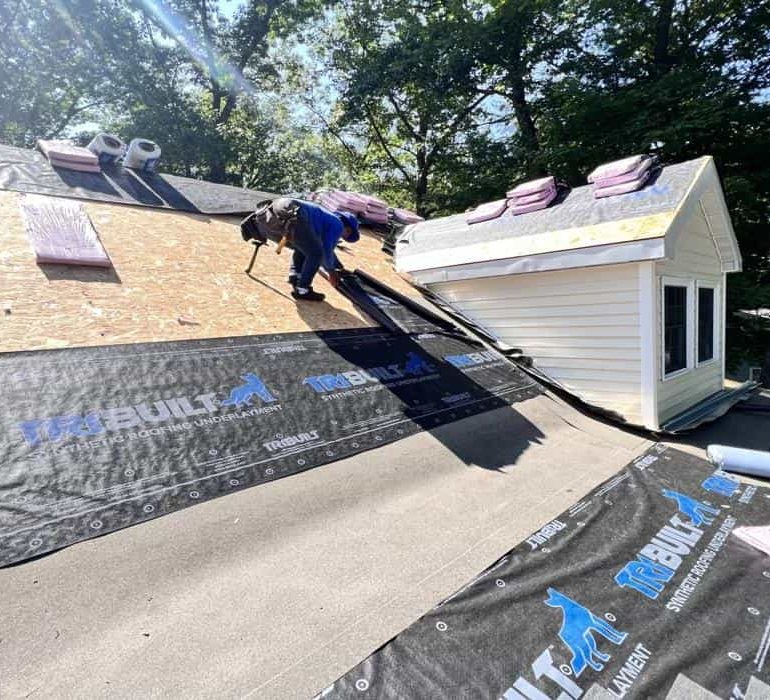 Professional roofing contractor installing synthetic roofing underlayment on residential house roof during roof replacement project