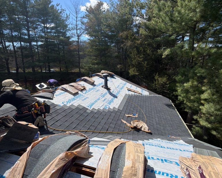 Asphalt shingle roof replacement in progress on a residential home surrounded by tall trees and wooded landscaping.